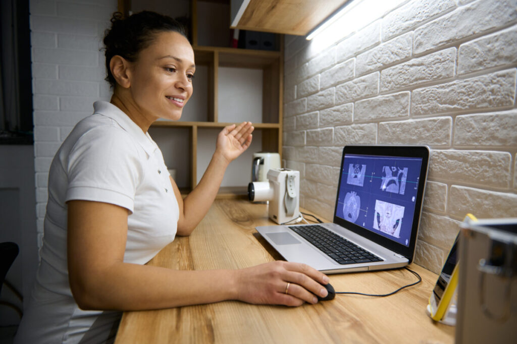 Woman doctor dentist sitting in front of a laptop with a panoramic x-ray of the human teeth and consulting a patient on a video call about the treatment. Online consultation concept in medical sphere