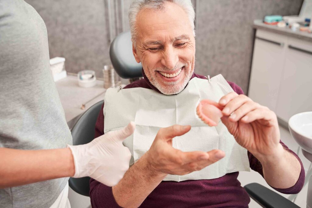 My new teeth. Cropped view of the dentist showing to senior grey haired patient teeth dentures while working at the dental clinic. Man sharing satisfaction smile